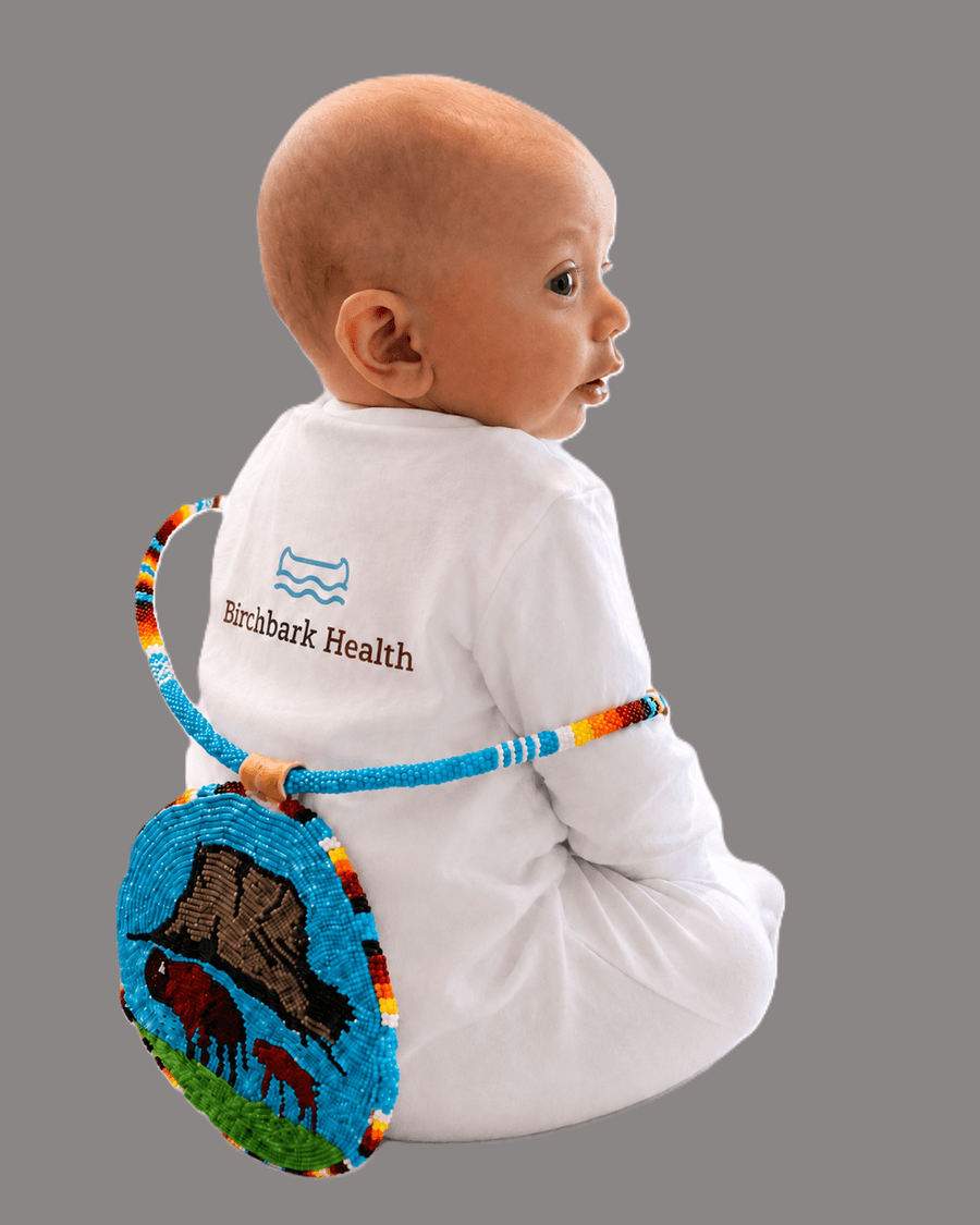 A baby in a Birchbark Health onesie wearing a beaded buffalo medallion