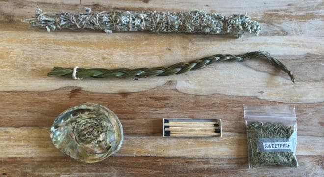 A smudge kit: sage, a sweetgrass braid, sweetpine, an abalone shell, and matches on a wooden table